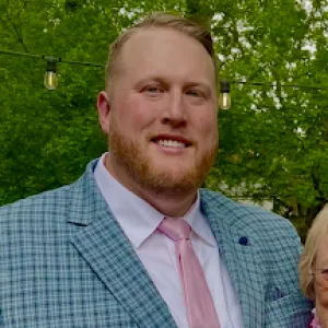A man smiling at the camera, wearing a light blue plaid blazer, white dress shirt, and light pink tie. He has short light-colored hair and a neatly trimmed beard.