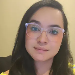 A woman in a yellow shirt in glasses smiling at the camera. She has dark brown hair and brown eyes.