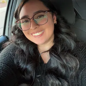 A woman with black hair and glasses smiling at the camera. She is wearing a black sweater and sitting in a vehicle.