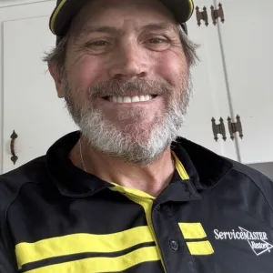 A man smiling at the camera, wearing a black and yellow ServiceMaster button up polo and hat.