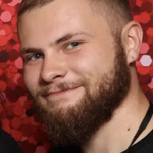 A man with a beard and buzzcut smiling at the camera. He is wearing a black shirt and has a red, sequined background behind him.