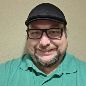 A bearded man smiling at the camera, standing in front of a wall. He is wearing a black hat, black glasses, and a light green polo.