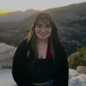 A woman with black and blonde hair smiling at the camera. She is on a mountaintop in a black cardigan.
